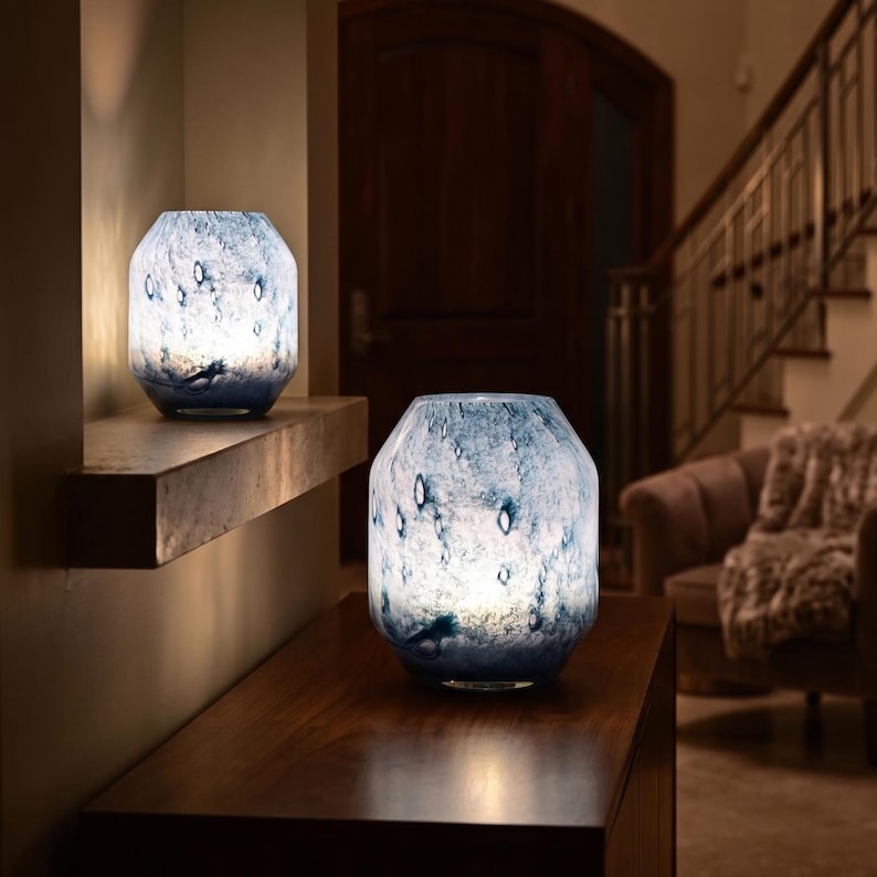 Puede incluir: Dos l&aacute;mparas de cristal iluminadas con un patr&oacute;n jaspeado de remolinos azul oscuro y blancos. Cada l&aacute;mpara tiene un cuerpo ancho y redondeado con la parte superior estrecha y una superficie lisa y transl&uacute;cida que emite un brillo c&aacute;lido.