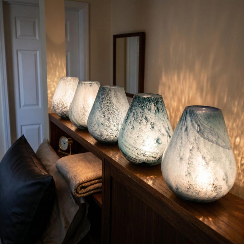 May include: Five teardrop-shaped glass lamps arranged in a row on a wooden surface. Each lamp features a unique mottled texture with varying shades of white, gray, and blue, emitting a soft, warm internal glow that illuminates the surrounding area.