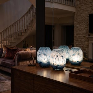 Puede incluir: Tres l&aacute;mparas cil&iacute;ndricas de cristal con un acabado moteado en azul y blanco. Las l&aacute;mparas presentan una superficie texturizada con patrones abstractos en azul oscuro y emiten un c&aacute;lido resplandor interno, sobre una superficie de madera.