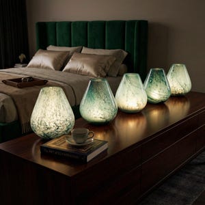 May include: Five teardrop-shaped glass lamps with a mottled green and white finish, each emitting a warm internal glow. The lamps are arranged in a row on a dark wooden surface next to a book titled The Art of Illumination and a small teacup.