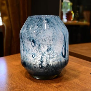 Puede incluir: Un florero cil&iacute;ndrico de vidrio con un acabado moteado en azul y blanco y una superficie lisa y brillante. El dise&ntilde;o muestra patrones de remolinos en tonos azul marino profundo y azul cielo m&aacute;s claro, creando un efecto marmolado en el vidrio.