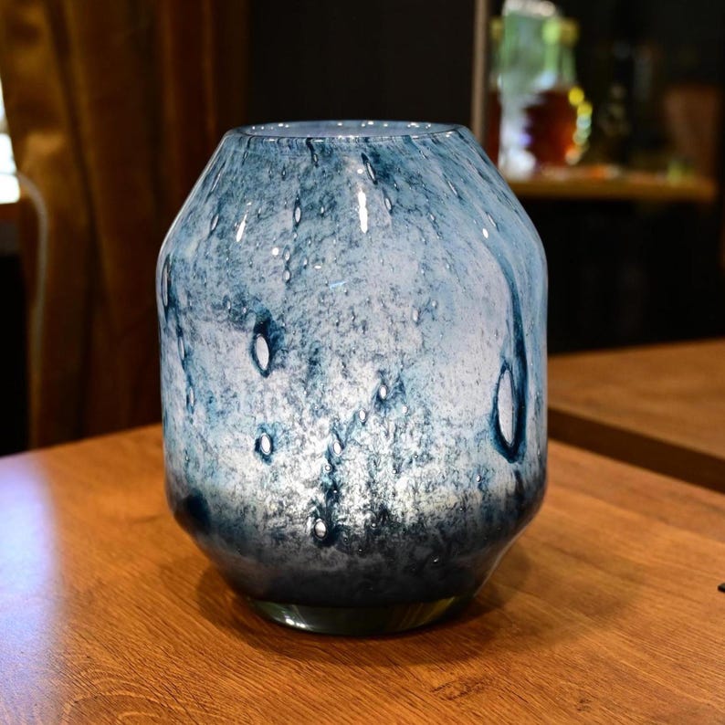 Puede incluir: Un florero cil&iacute;ndrico de vidrio con un acabado moteado en azul y blanco y una superficie lisa y brillante. El dise&ntilde;o muestra patrones de remolinos en tonos azul marino profundo y azul cielo m&aacute;s claro, creando un efecto marmolado en el vidrio.