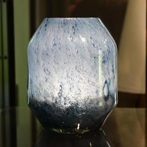 Puede incluir: Un florero de vidrio con forma redondeada y c&oacute;nica que presenta un patr&oacute;n moteado que transiciona desde un azul y blanco claro y transl&uacute;cido en la parte superior hasta un azul marino profundo y s&oacute;lido en la base, con una superficie pulida.