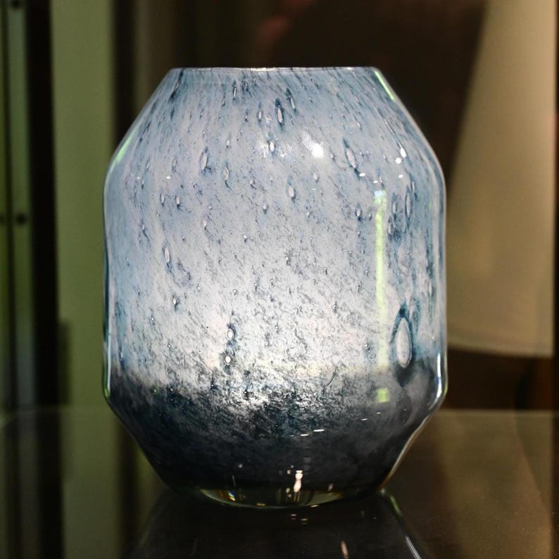 Puede incluir: Un florero de vidrio con forma redondeada y c&oacute;nica que presenta un patr&oacute;n moteado que transiciona desde un azul y blanco claro y transl&uacute;cido en la parte superior hasta un azul marino profundo y s&oacute;lido en la base, con una superficie pulida.