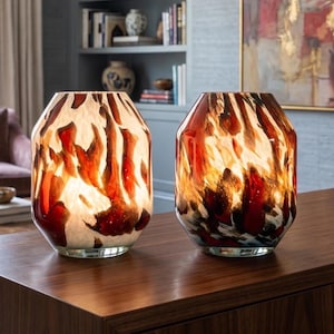May include: Two glass table lamps with a cylindrical shape and a wide opening at the top. The glass features a marbled pattern with swirling colors of deep red, burnt orange, black, and white, creating a flame-like appearance when illuminated.