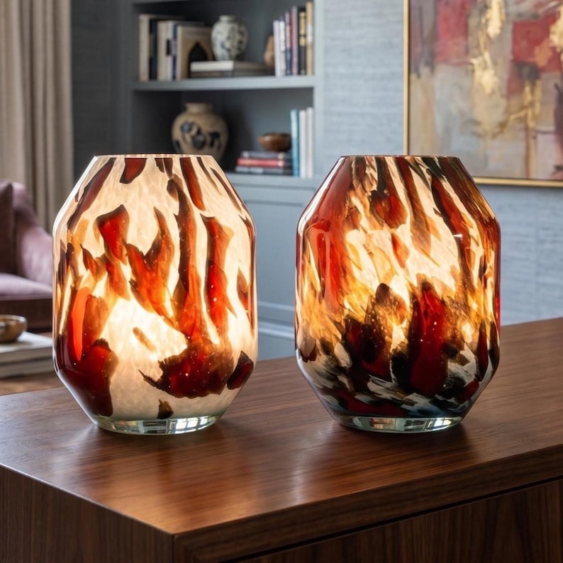 May include: Two glass table lamps with a cylindrical shape and a wide opening at the top. The glass features a marbled pattern with swirling colors of deep red, burnt orange, black, and white, creating a flame-like appearance when illuminated.