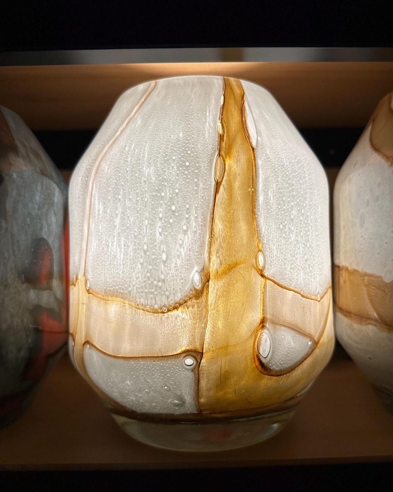 May include: A glass vase featuring a rounded, tapered silhouette with a textured white surface containing numerous small air bubbles. The piece is accented by vertical and horizontal bands of translucent amber-colored glass swirls.