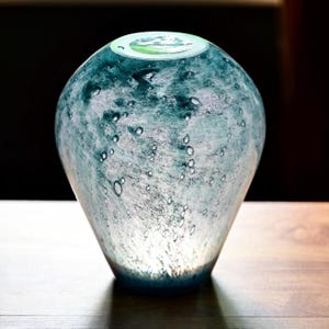 Puede incluir: Un jarr&oacute;n de vidrio en forma de l&aacute;grima con una paleta de colores verde azulado y blanco moteado con numerosas burbujas de aire incrustadas en todo el cuerpo. El jarr&oacute;n tiene un exterior liso y redondeado y una abertura circular superior.