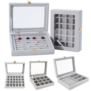 Puede incluir: Una colección de joyeros de terciopelo gris con tapas de vidrio transparente y cierres dorados. Las cajas están abiertas, revelando ranuras para anillos y compartimentos. Algunas cajas están vacías, mientras que otras exhiben anillos con acentos coloridos.