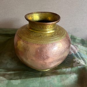 May include: A copper and brass pot with a wide, rounded body and a narrow neck. The upper section is brass with etched floral patterns, while the lower body is copper. The pot sits on a green and white floral patterned fabric.