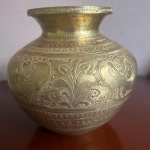 Puede incluir: Un pequeño jarrón antiguo de latón con cuerpo redondeado y borde acampanado. El jarrón está adornado con intrincados diseños florales y foliares grabados. El latón tiene un cálido tono dorado y el jarrón parece bien conservado.