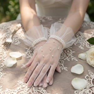 May include: Sheer white gloves with pearl accents. The gloves feature a delicate, ruffled cuff adorned with multiple rows of pearls. Small pearls are also scattered across the glove's surface. The gloves are likely for a special occasion, such as a wedding.