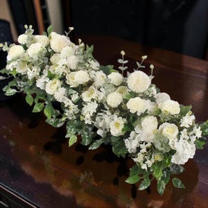 Könnte beinhalten: Ein üppiges Blumenarrangement mit weißen Rosen, Pomponblüten und grünem Laub. Die Anordnung befindet sich auf einer dunklen Holzoberfläche, was einen schönen Kontrast erzeugt.