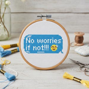 May include: A cross-stitch embroidery hoop with the text "No worries if not!!!" in blue and white. The design includes a yellow emoji. The wooden hoop is surrounded by craft supplies, including thread and scissors.