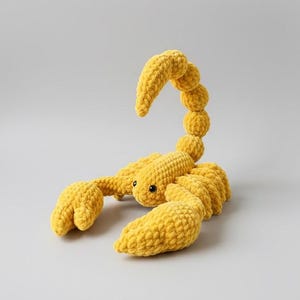 May include: A handmade, yellow crochet scorpion toy. The scorpion has a segmented body, large claws, and a curved tail. The toy is made of soft, textured yarn and features black safety eyes.