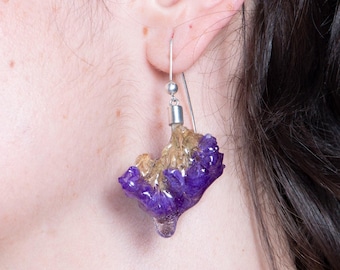 May include: A close-up of a person wearing a pair of amethyst crystal earrings. The earrings are a cluster of purple and gold crystals, suspended from silver hooks. The earrings are a unique and natural accessory.