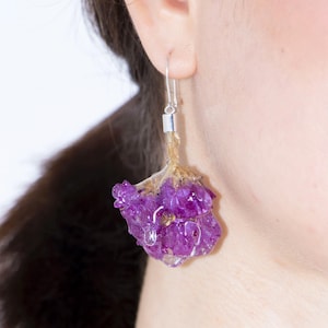 May include: A single earring featuring a vibrant purple flower encased in clear resin. The earring hangs from a silver hook and post. The flower has a natural, organic shape, with visible details and textures. The earring is worn on a person's ear.