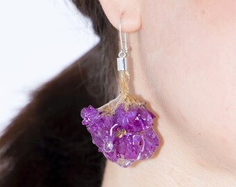 May include: A single earring featuring a vibrant purple flower encased in clear resin. The earring hangs from a silver hook and post. The flower has a natural, organic shape, with visible details and textures. The earring is worn on a person's ear.