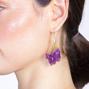 May include: A close-up of a person's ear wearing a pair of unique earrings. The earrings feature a silver hook, a translucent yellow-brown section, and a cluster of deep purple, textured material, resembling a flower or gemstone.