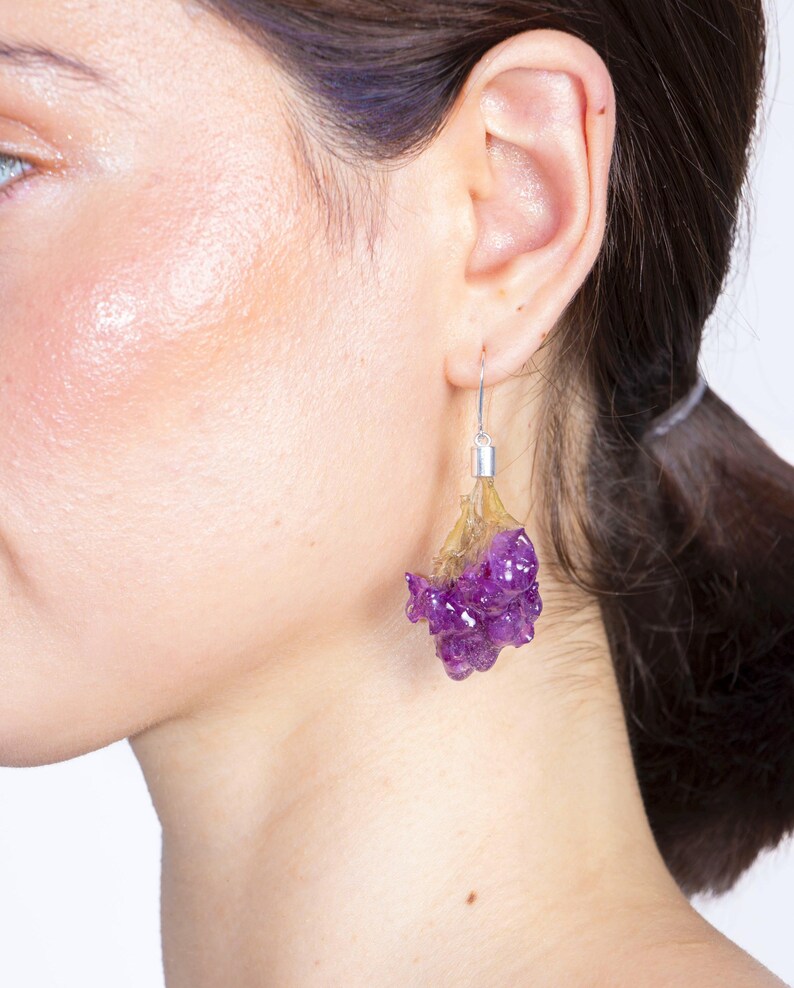 May include: A close-up of a person's ear wearing a pair of unique earrings. The earrings feature a silver hook, a translucent yellow-brown section, and a cluster of deep purple, textured material, resembling a flower or gemstone.