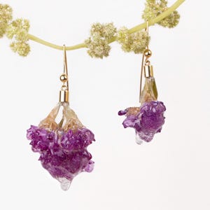 May include: A pair of gold-toned earrings featuring purple flower buds encased in clear resin. The earrings hang from delicate gold hooks, with a light-colored stem and small green buds in the background. The earrings are a unique floral accessory.
