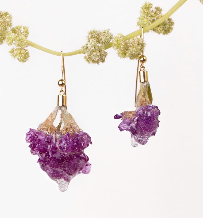 May include: A pair of gold-toned earrings featuring purple flower buds encased in clear resin. The earrings hang from delicate gold hooks, with a light-colored stem and small green buds in the background. The earrings are a unique floral accessory.
