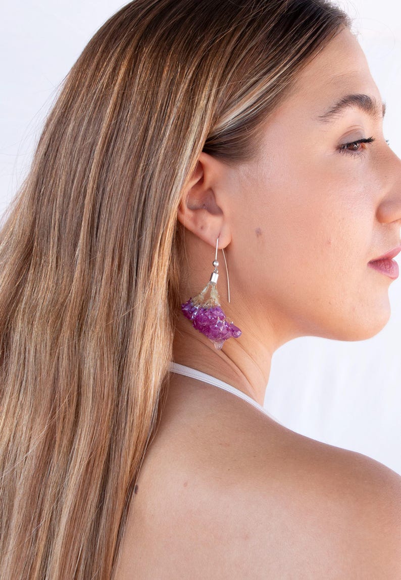May include: A close-up of a woman wearing a pair of unique earrings. The earrings feature a silver hook with a cluster of purple and clear crystal-like material. The woman has long, light brown hair.