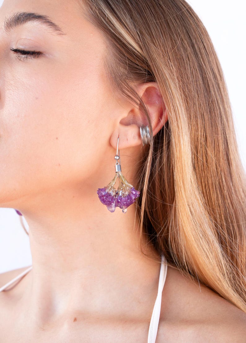 May include: A close-up of a person wearing a pair of silver drop earrings. Each earring features a cluster of small, vibrant purple crystals, suspended from a silver hook. The earrings have a delicate, floral appearance.