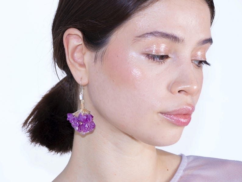 May include: A close-up of a person wearing a silver earring with a small rectangular accent and a cluster of purple flowers. The earring is dangling from the ear. The person has dark hair and is looking down.
