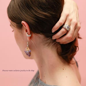 May include: A person wearing a silver ring with a purple stone and a silver earring with a cluster of purple crystals. The image is set against a pink background. The text in the image says, "Discover more exclusive jewelry in the shop."