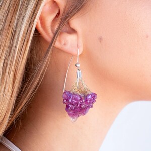 May include: A close-up of a dangling earring with a silver hook. The earring features a cluster of purple and clear resin, resembling a flower. The resin is suspended from a silver post, creating a unique and artistic accessory.