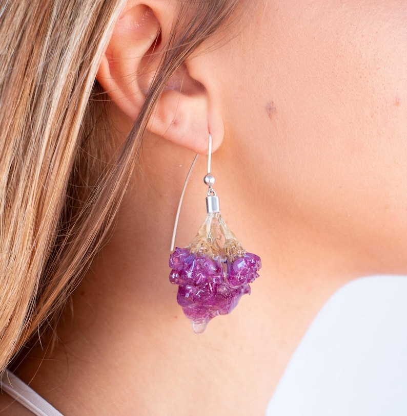 May include: A close-up of a dangling earring with a silver hook. The earring features a cluster of purple and clear resin, resembling a flower. The resin is suspended from a silver post, creating a unique and artistic accessory.