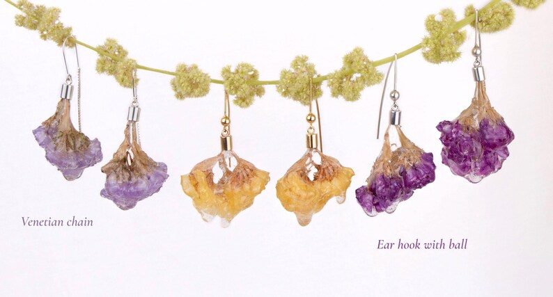 May include: A collection of handmade floral earrings. The earrings feature real pressed flowers in shades of purple and yellow, suspended from silver and gold-colored ear hooks. Some earrings have a Venetian chain.