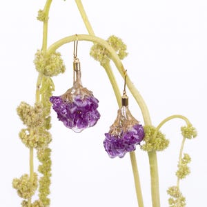 May include: A pair of gold-toned drop earrings with purple, flower-shaped pendants. The earrings are suspended from delicate gold hooks and are set against a backdrop of light green stems and small, yellow-green flower clusters.