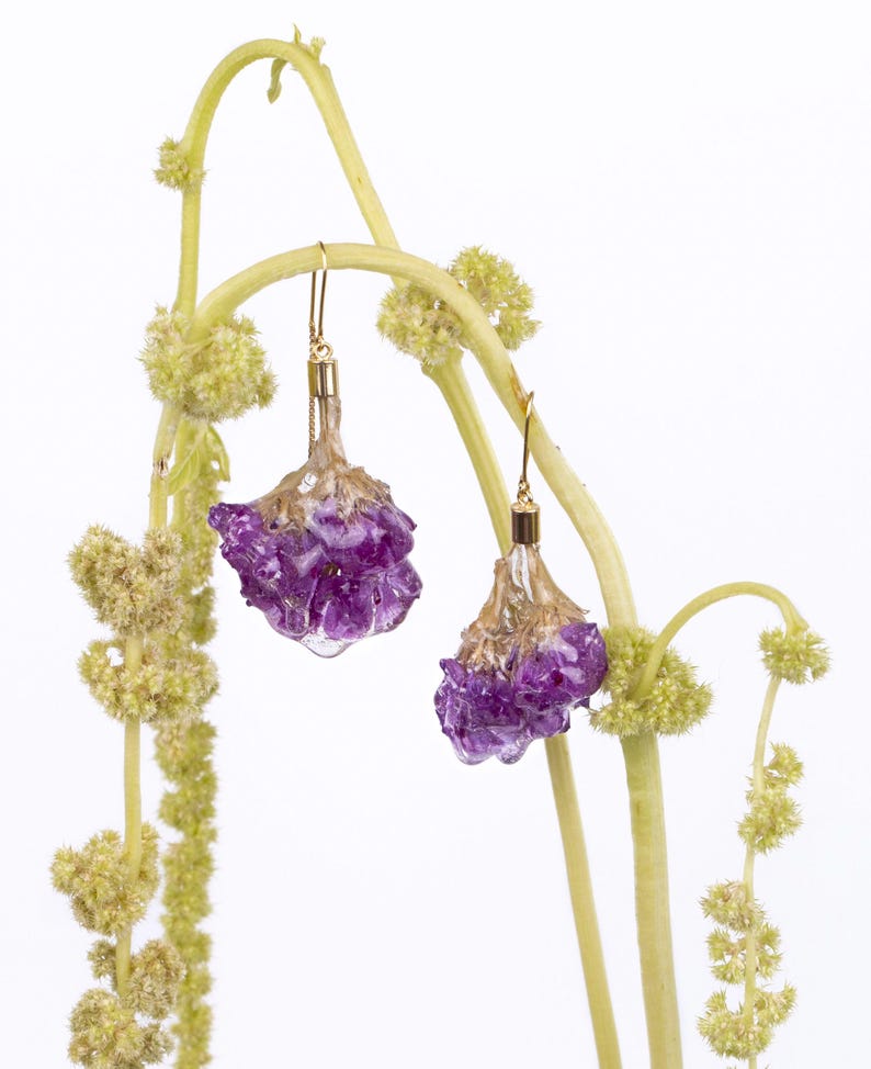 May include: A pair of gold-toned drop earrings with purple, flower-shaped pendants. The earrings are suspended from delicate gold hooks and are set against a backdrop of light green stems and small, yellow-green flower clusters.