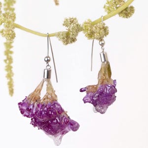 May include: A pair of handmade earrings featuring vibrant purple flowers encased in clear resin. The earrings hang from silver-colored hooks, with a small silver bead and a cylindrical silver component. The flowers have a natural, organic appearance.