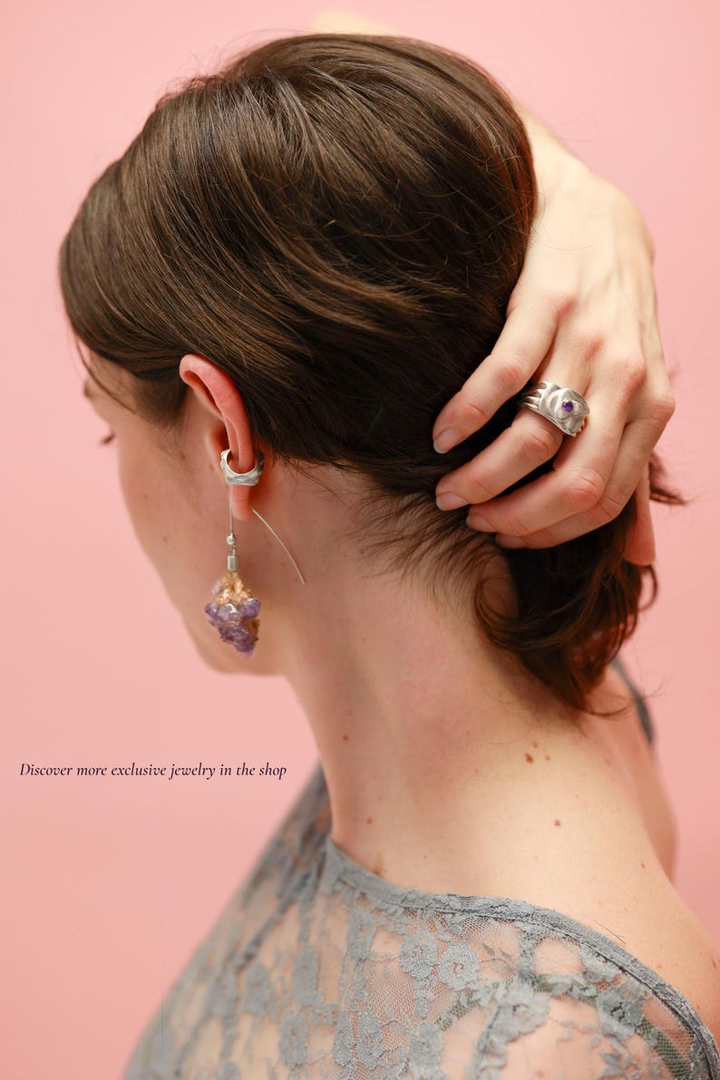 May include: A person wearing a silver ring with a purple stone and a silver earring with a cluster of purple crystals. The image is set against a pink background. The text in the image says, "Discover more exclusive jewelry in the shop."