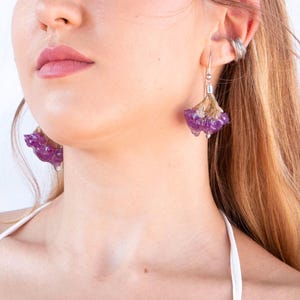 May include: Close-up of a person wearing unique earrings. The earrings feature a purple, flower-like design with a silver hook. The earrings are a striking accessory, adding a touch of nature-inspired elegance.