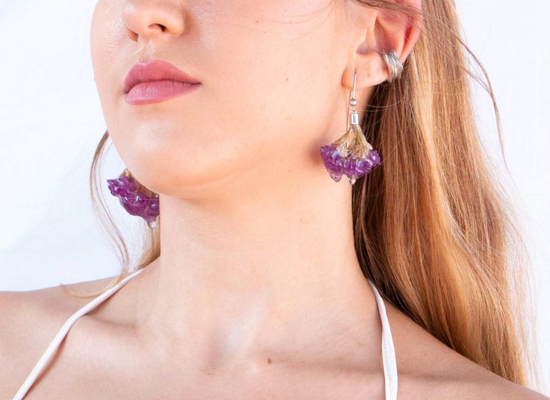May include: Close-up of a person wearing unique earrings. The earrings feature a purple, flower-like design with a silver hook. The earrings are a striking accessory, adding a touch of nature-inspired elegance.
