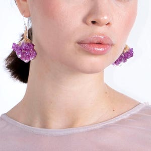 May include: Close-up of amethyst earrings. The earrings feature clusters of purple crystals with a gold-colored base, suspended from silver-colored hooks. The earrings are worn by a person with fair skin and a light pink top.
