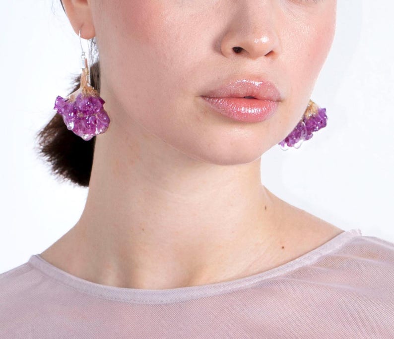 May include: Close-up of amethyst earrings. The earrings feature clusters of purple crystals with a gold-colored base, suspended from silver-colored hooks. The earrings are worn by a person with fair skin and a light pink top.