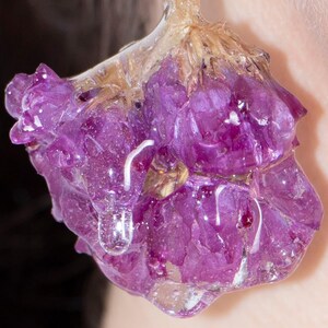 May include: Close-up of a purple flower encased in clear resin. The flower has multiple petals and is a vibrant shade of purple. The resin has a glossy, wet appearance, with a few air bubbles visible. The flower is suspended in the resin, creating a unique, artistic piece.