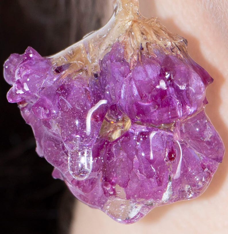 May include: Close-up of a purple flower encased in clear resin. The flower has multiple petals and is a vibrant shade of purple. The resin has a glossy, wet appearance, with a few air bubbles visible. The flower is suspended in the resin, creating a unique, artistic piece.