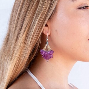 May include: A close-up of a silver earring with a cluster of purple beads. The earring is attached to a silver hook. The beads are clustered together, creating a unique and eye-catching design. The earring is worn by a person with blonde hair.