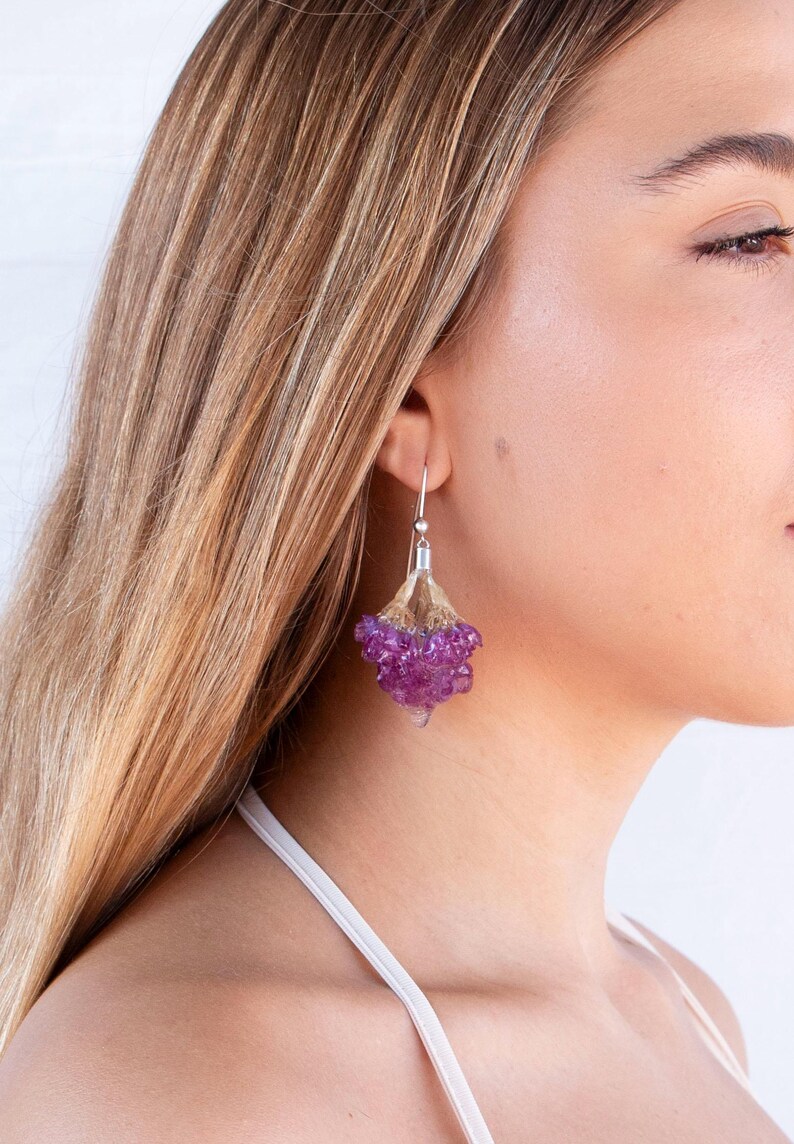 May include: A close-up of a silver earring with a cluster of purple beads. The earring is attached to a silver hook. The beads are clustered together, creating a unique and eye-catching design. The earring is worn by a person with blonde hair.