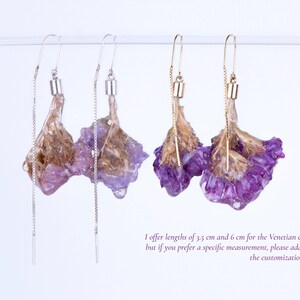 May include: Earrings featuring dried purple flowers encased in clear resin. The earrings have a delicate chain and hook design. One pair has silver-colored hooks and chains, while the other has gold-colored hooks and chains.