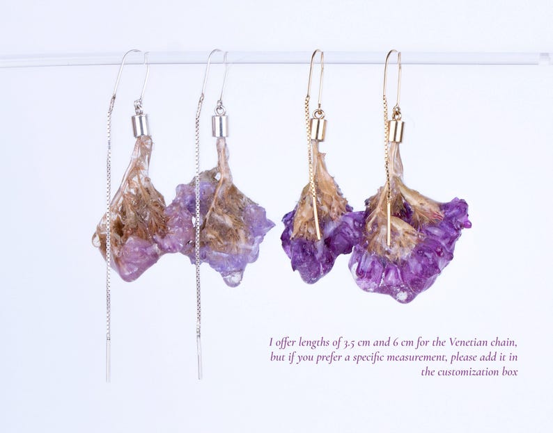 May include: Earrings featuring dried purple flowers encased in clear resin. The earrings have a delicate chain and hook design. One pair has silver-colored hooks and chains, while the other has gold-colored hooks and chains.