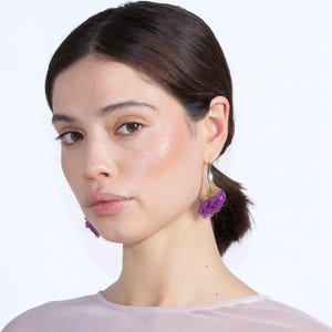 May include: A woman wearing purple and gold earrings. The earrings have a cluster design with a silver hook. The earrings are a vibrant purple color with gold accents.