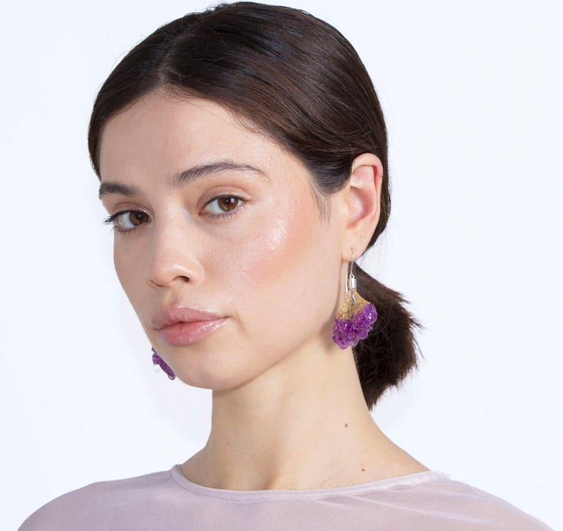 May include: A woman wearing purple and gold earrings. The earrings have a cluster design with a silver hook. The earrings are a vibrant purple color with gold accents.