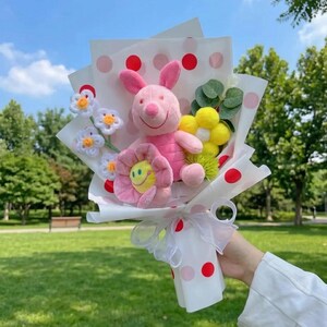 K&ouml;nnte beinhalten: Ein Blumenstrau&szlig; mit einem rosa Pl&uuml;schferkel, einer gelben Pl&uuml;schblume mit Smiley-Gesicht und mehreren kleinen wei&szlig;en geh&auml;kelten Blumen mit gelben Zentren. Das Arrangement ist in wei&szlig;es Papier mit roten Punkten und ein wei&szlig;es Schleifenband gewickelt.